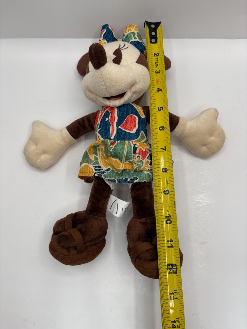 May include: A Minnie Mouse plush doll, approximately 12 inches tall, wearing a colorful dress and bow. The doll has brown ears, arms, and legs, with beige hands and face. The dress features a blue, red, and green tropical pattern. The doll is wearing brown sandals.