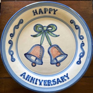 May include: A round ceramic plate with a cream-colored center and a blue rim. The plate features a hand-painted design of two bells tied with a green ribbon, flanked by blue scroll patterns. The text HAPPY is at the top and ANNIVERSARY at the bottom.