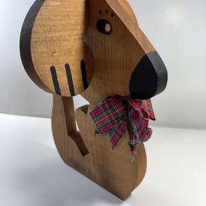 May include: Wooden dog sculpture with a black nose, eye, and ear markings. The dog wears a red, green, and black plaid ribbon around its neck. The sculpture is made of light brown wood.