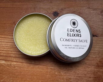 May include: An open, round, silver tin of Comfrey Salve by Edens Elixirs, with the lid beside it. The salve is a light yellow color and has a slightly textured appearance. The lid has a white label with black text and a sunburst logo.