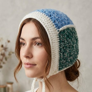 Peut inclure: Un bonnet en crochet présentant un design color-block avec un panneau supérieur bleu clair et des panneaux latéraux vert foncé, le tout bordé d'une épaisse bordure crème. Le bonnet comprend deux liens tressés crème à nouer sous le menton.