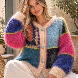 Peut inclure: Un cardigan à manches longues en maille duveteuse avec un motif patchwork composé de blocs bleus, roses, orange et noirs. Le vêtement présente un col en V, une bordure beige côtelée et trois boutons décoratifs carrés bleu clair.