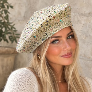 Op de afbeelding: Een textuurrijke baret van lichtbeige, met goudkleurige metaaldraad doorweven breiwerk, versierd met kleine, verspreide veelkleurige lovertjes in blauw, roze, groen en zilver, voorzien van een dikke, geribbelde gebreide boord aan de onderkant.