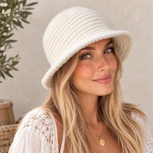 Peut inclure: Un chapeau bob de couleur crème présentant un motif en maille crochetée côtelée et texturée. Le chapeau possède une calotte arrondie et un bord incliné vers le bas, fabriqué à partir d'un fil doux avec un point uniforme et régulier.