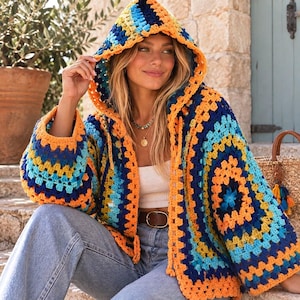 Op de afbeelding: Een vest met capuchon voorzien van een levendig granny square haakpatroon in tinten oranje, geel, lichtblauw en donkerblauw. Het kledingstuk heeft lange mouwen, een open voorzijde en een capuchon, gemaakt van garen met een textuurrijk breipatroon.