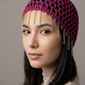 May include: A magenta crochet head covering with a lattice design. Gold-colored chain fringe hangs from the front. The head covering is worn by a person with dark hair and a neutral background.