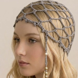 May include: A gray crocheted head covering with a net-like design. A strand of the same gray yarn hangs down, adorned with a pearl and three gold-colored beads. The head covering is worn on a person with blonde hair.