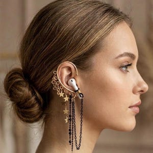 Peut inclure: Un ear cuff doré avec un écouteur blanc, orné d'étoiles dorées pendantes et de chaînes de perles noires. L'ear cuff s'enroule autour de l'oreille, créant un accessoire élégant et unique. Le design allie fonctionnalité et mode.