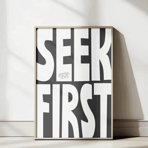 May include: A framed print featuring the words "SEEK FIRST" in large, white, bold letters on a dark grey background. A smaller line of text is visible at the top. The frame is a light wood colour, and the artwork is displayed on a white wall.