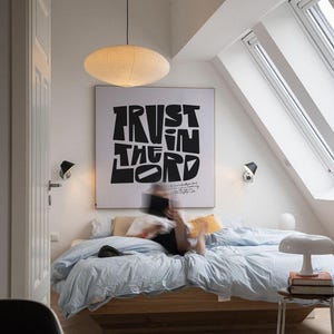 Op de afbeelding: Een slaapkamer met een wit plafond en witte muren. Een groot raam met een wit kozijn bevindt zich in de rechterbovenhoek van de afbeelding. Een bed met een wit hoofdeinde en een lichtblauwe deken staat in het midden van de afbeelding. Een persoon ligt op het bed. Een zwart-witte poster met de tekst "TRUST IN THE LORD" hangt aan de muur boven het bed. Een witte lamp staat op een klein tafeltje naast het bed.