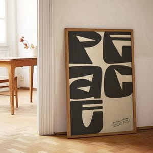 May include: A rectangular framed wall art piece featuring large, bold, black abstract typography that spells out the word PEACE against a light beige background. The frame is made of light-toned wood, and small cursive text is visible in the bottom right corner.