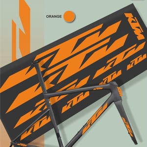 May include: A dark gray bicycle frame with orange KTM logos and accents. The frame is positioned in front of a black background with repeating orange KTM logos. The word "ORANGE" is above an orange circle.
