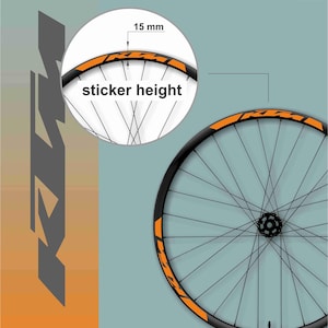 May include: A bicycle wheel with a black rim and orange accents. The wheel has thin black spokes and a central hub. The image includes a close-up showing the sticker height of 15 mm. The background is a two-tone teal and orange.