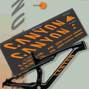 May include: Black mountain bike frame with orange "Canyon" lettering and a sheet of orange decals. The decal sheet includes the text "Canyon", "Fox", "Rock Shox", "Shimano", and "SRAM". The dimensions of the decals are 300 x 40 mm, 61 x 30 mm, and 20 x 10 mm.