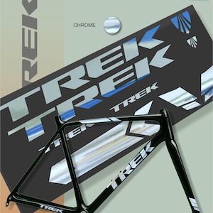 Könnte beinhalten: Ein schwarzer Trek-Fahrradrahmen mit weißen und silbernen Trek-Logos und Akzenten. Das Bild enthält auch einen Bogen mit Trek-Aufklebern mit Chrom- und blauen Details sowie das Wort "Chrome". Der Hintergrund ist ein Farbverlauf aus Grün und Beige.
