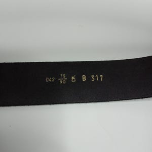 May include: Close-up of a black leather belt with gold lettering. The belt has the following measurements: D42, 75/90, and B 317. The belt is on a white background.