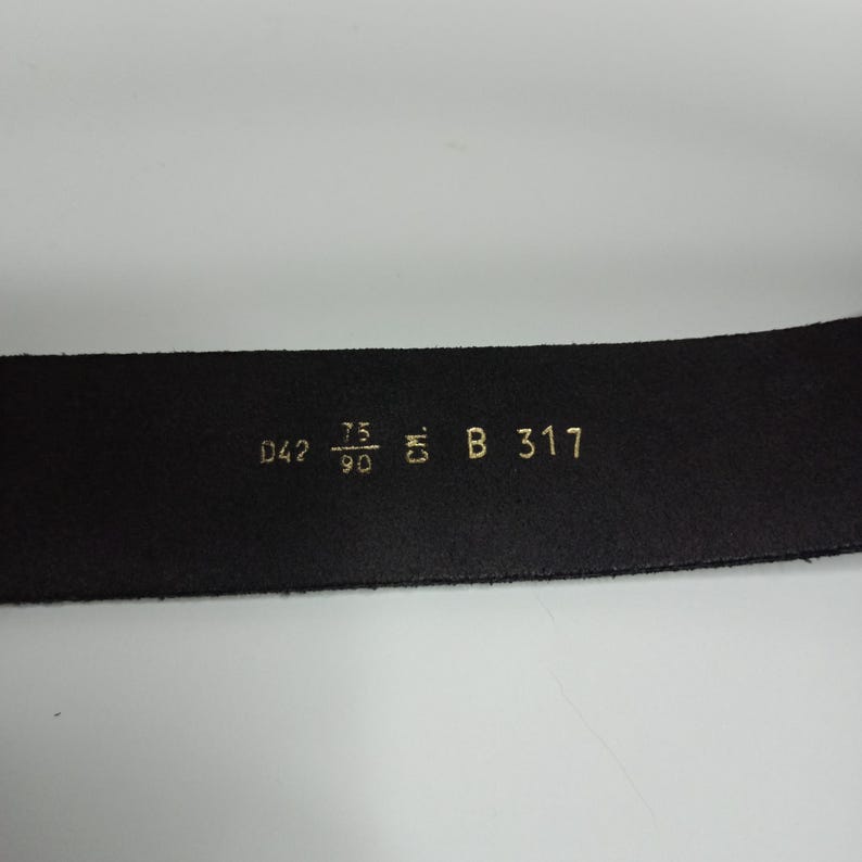 May include: Close-up of a black leather belt with gold lettering. The belt has the following measurements: D42, 75/90, and B 317. The belt is on a white background.