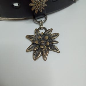 May include: A black leather collar with a silver-toned flower pendant. The pendant is a detailed, multi-petaled flower with a tarnished finish. Another flower adorns the collar. The collar has a silver-toned ring and a buckle.