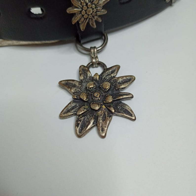 May include: A black leather collar with a silver-toned flower pendant. The pendant is a detailed, multi-petaled flower with a tarnished finish. Another flower adorns the collar. The collar has a silver-toned ring and a buckle.