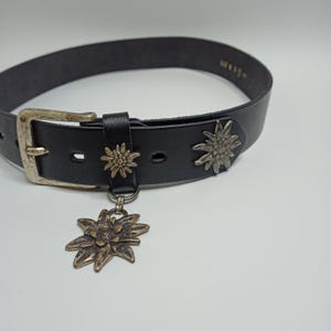 May include: Black leather belt with a silver-colored buckle and decorative metal flower accents. A larger flower charm hangs from the belt. The belt is a classic accessory.