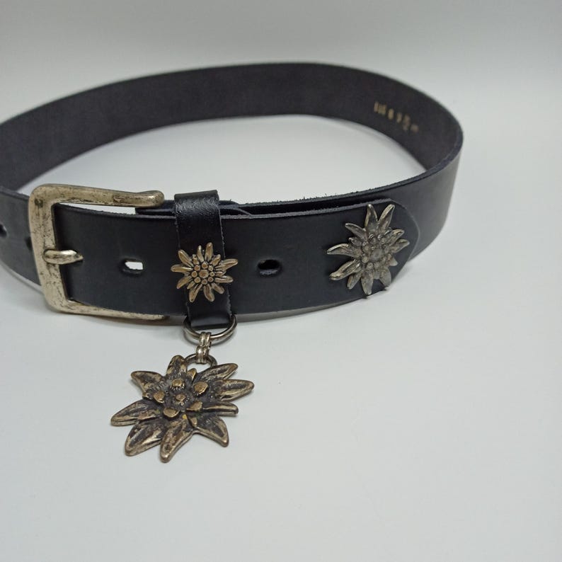 May include: Black leather belt with a silver-colored buckle and decorative metal flower accents. A larger flower charm hangs from the belt. The belt is a classic accessory.
