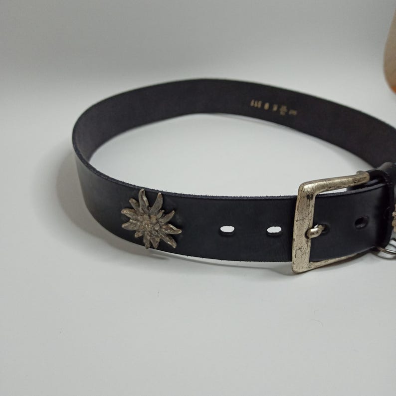 May include: A black leather belt with a silver buckle and a decorative metal flower. The belt has a classic design with a single loop and multiple holes for adjustment. The metal flower is a prominent detail.