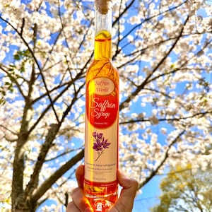 Peut inclure: Une bouteille en verre transparente remplie de sirop de safran, tenue devant un fond d'arbre en fleurs et de ciel bleu. L'étiquette indique "Saffron World Saffron Syrup Charlotte NC". La bouteille contient 175 ml de sirop.