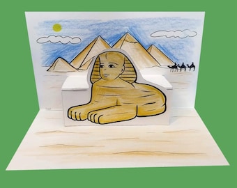 May include: A pop-up card featuring a hand-drawn illustration of the Sphinx and pyramids in Egypt. The Sphinx is a light brown color, and the pyramids are a tan color. The background includes a blue sky, a yellow sun, and a few clouds.