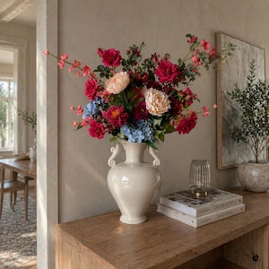 Op de afbeelding: Een crèmekleurige vaas met een groot bloemstuk van rode, roze, perzik- en blauwe bloemen. De vaas staat op een houten console met boeken en een decoratief glazen object. De achtergrond is een neutraal getinte muur.