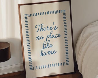 May include: A framed print with the text "There's no place like home" in blue script. A black lamp and a coffee cup on a book with red and pink flowers are on the wooden floor. A bed with white linens is in the background.