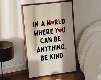 May include: A framed art print with the text "IN A WORLD WHERE YOU CAN BE ANYTHING, BE KIND" in black font with a red heart. A coffee cup sits on a book with colorful flowers. A black lamp is on the left.