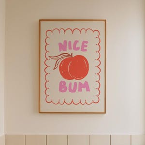 Könnte beinhalten: Ein gerahmter Kunstdruck in einem Badezimmer. Der Druck zeigt eine Pfirsich-Illustration mit den Worten "NICE BUM" in Rosa. Das Badezimmer hat eine weiße Badewanne, einen Holzschemel und einen schwarz-weiß karierten Boden.