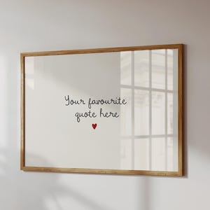 May include: A framed print with a light wood frame hangs on a white wall. The print features the text "Your favourite quote here" in a handwritten style, with a small red heart below the text. The frame is rectangular.