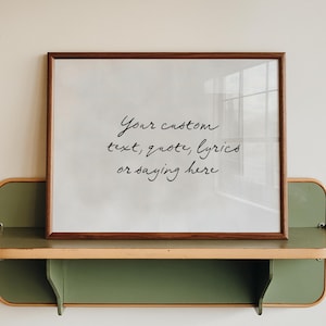 Op de afbeelding: Een ingelijste print met de tekst "Your custom text, quote, lyrics or saying here." De lijst is bruin en de print wordt weergegeven op een groen en beige plankje.