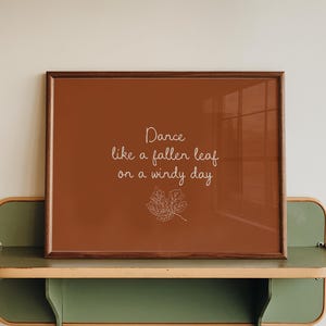 May include: A rectangular wall art print in a dark brown frame featuring a burnt orange background with the white text "Dance like a fallen leaf on a windy day" and a simple white line drawing of a single leaf centered below the text.