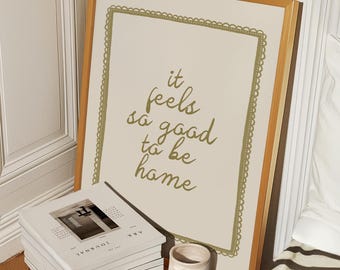 May include: A framed print with a gold frame and a white background. The print features the text "it feels so good to be home" in a gold script font.
