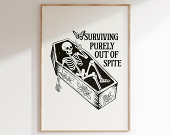 May include: Black and white framed art print featuring a skeleton in a coffin with the text "SURVIVING PURELY OUT OF SPITE." The coffin is decorated with floral designs and a butterfly.