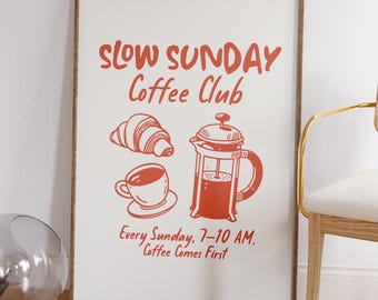 May include: Framed print with a coffee theme. The print features the words "Slow Sunday Coffee Club" in red, along with illustrations of a croissant, coffee cup, and French press. The text "Every Sunday, 7-10 AM. Coffee Comes First" is also included.