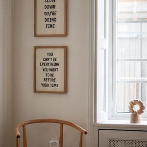 May include: Two framed prints with inspirational quotes hang on a cream-colored wall. The top print reads "SLOW DOWN YOU'RE DOING FINE." The bottom print says "YOU CAN'T BE EVERYTHING YOU WANT TO BE BEFORE YOUR TIME."