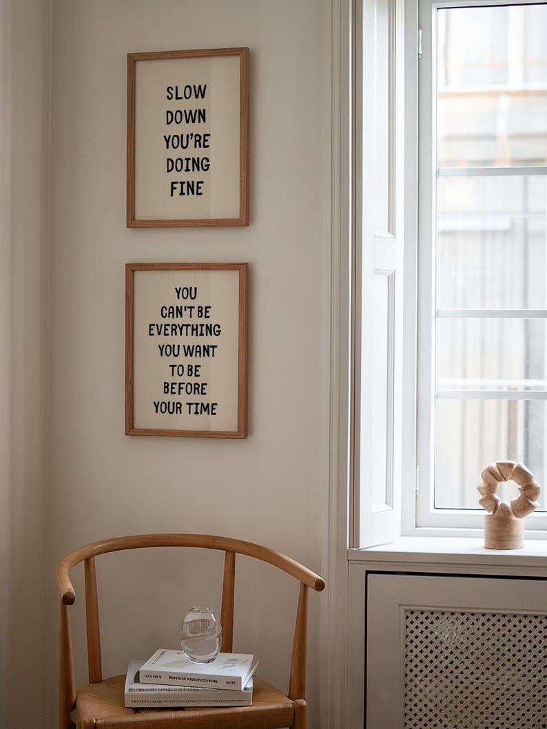 May include: Two framed prints with inspirational quotes hang on a cream-colored wall. The top print reads "SLOW DOWN YOU'RE DOING FINE." The bottom print says "YOU CAN'T BE EVERYTHING YOU WANT TO BE BEFORE YOUR TIME."