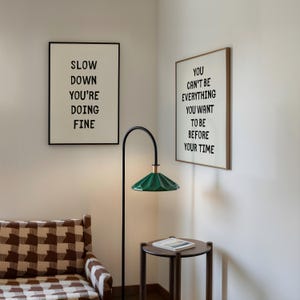 May include: Interior scene with two framed prints with black text on a white background. One says "SLOW DOWN YOU'RE DOING FINE" and the other says "YOU CAN'T BE EVERYTHING YOU WANT TO BE BEFORE YOUR TIME". A green lamp and a small table are also visible.