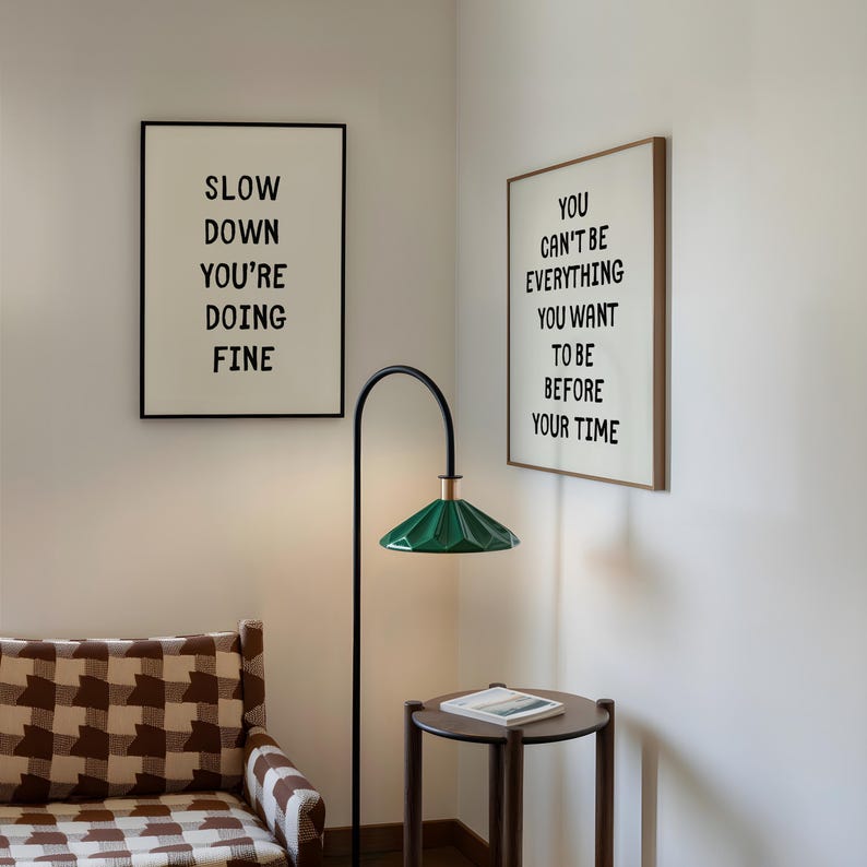 May include: Interior scene with two framed prints with black text on a white background. One says "SLOW DOWN YOU'RE DOING FINE" and the other says "YOU CAN'T BE EVERYTHING YOU WANT TO BE BEFORE YOUR TIME". A green lamp and a small table are also visible.