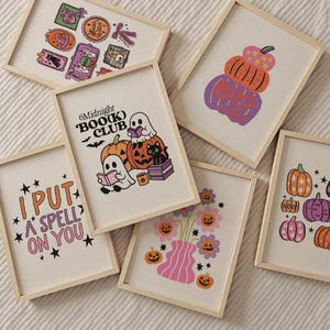 May include: A collection of framed Halloween-themed art prints. The prints feature illustrations of pumpkins, ghosts, and floral arrangements in a retro style. One print reads "I PUT A SPELL ON YOU," and another says "Midnight Book Club."