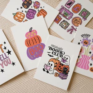 May include: A collection of Halloween-themed greeting cards featuring colorful illustrations. Designs include pumpkins, ghosts, cats, and the text "Midnight BOO(k) Club" and "Spooky in Bloom". The cards are on a neutral background.