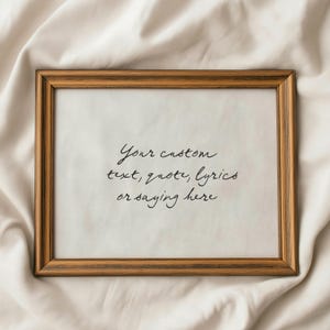 May include: A rectangular wooden picture frame with a light brown finish. The frame holds a white paper with the handwritten text: "Your custom text, quote, lyrics or saying here."