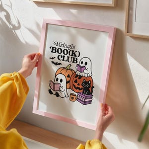 May include: A pink framed print featuring the text "Midnight BOO(K) CLUB" with illustrations of ghosts, pumpkins, a black cat, and books. The artwork has a Halloween theme with a cartoon style.