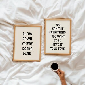 May include: Two framed prints with inspirational quotes on a white background. One reads "SLOW DOWN YOU'RE DOING FINE" and the other says "YOU CAN'T BE EVERYTHING YOU WANT TO BE BEFORE YOUR TIME". A cup of coffee is held in the hand.