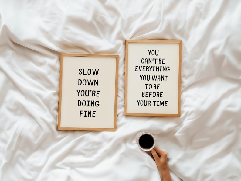 May include: Two framed prints with inspirational quotes on a white background. One reads "SLOW DOWN YOU'RE DOING FINE" and the other says "YOU CAN'T BE EVERYTHING YOU WANT TO BE BEFORE YOUR TIME". A cup of coffee is held in the hand.