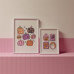 May include: Two framed Halloween-themed art prints. The larger print features illustrated pumpkins in orange, pink, and purple with star accents. The smaller print showcases a pumpkin, bats, and a ghost. Both prints are in white frames.