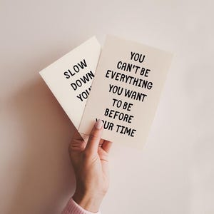 May include: Two cream-colored cards with black text are held in a hand. One card reads "SLOW DOWN YOU" and the other reads "YOU CAN'T BE EVERYTHING YOU WANT TO BE BEFORE YOUR TIME."